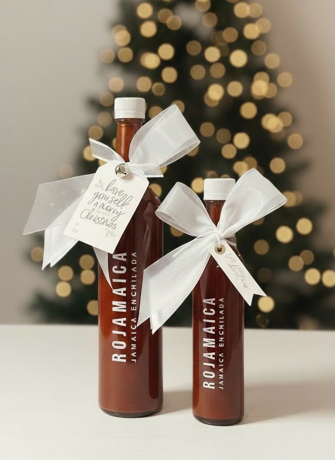 Two bottles of 'Rojamica' sauce with ribbons in front of a Christmas tree.